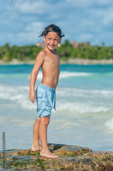 Obraz Niño en traje de baño azul sonríe parado sobre roca en las aguas azules del Mar Caribe.