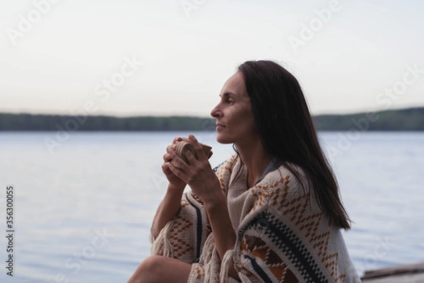 Obraz A peaceful beautiful dark haired young woman sits by the lake with a сup of tea and looks into the distance with pleasure