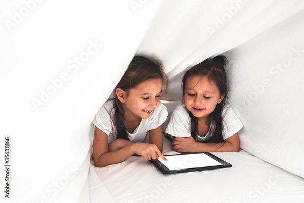 Fototapeta Two beautiful little sisters lying in the bed and look at the screen of a tablet, smart technology