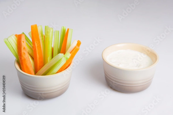 Fototapeta close up isolated shot of a mixed bowl of crunchy orange carrot slices and juicy green celery sticks with a white cup of blue cheese dipping sauce on a white background