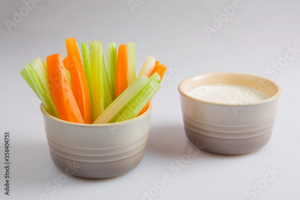 Fototapeta close up isolated shot of a mixed bowl of crunchy orange carrot slices and juicy green celery sticks with a white cup of blue cheese dipping sauce on a white background