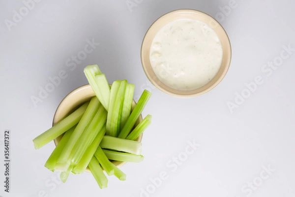 Fototapeta close up isolated flat lay top view shot of a bowl of crunchy juicy green celery sticks next to a white cup of blue cheese dipping sauce on a white background