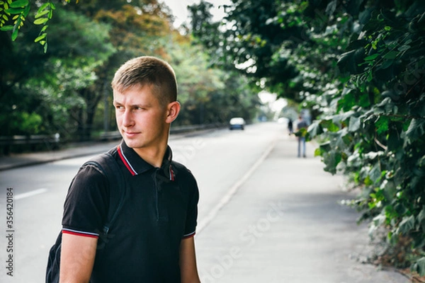 Obraz Portrait of a young man in a black T-shirt looking back on the background of a road and green trees.