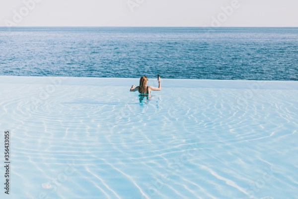 Fototapeta Girl takes pictures on the phone by the ocean while standing in the hotel’s infinity pool. Long hair, bikini.
