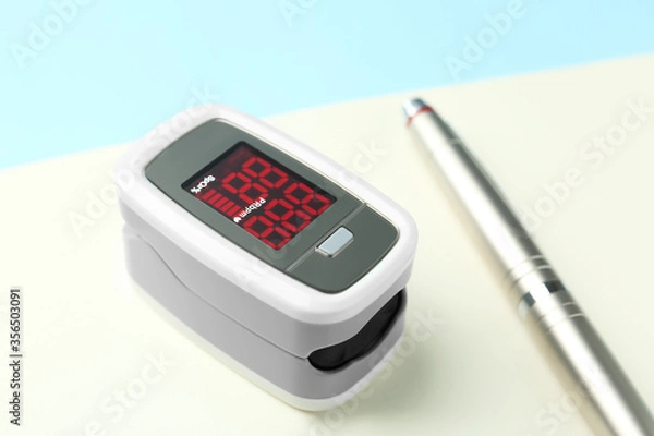 Fototapeta Modern fingertip pulse oximeter, pen and open notebook on light blue background, closeup