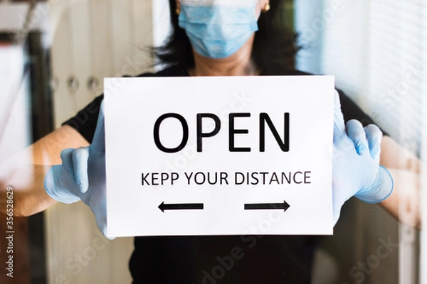 Fototapeta woman puts an "open, keep your distance" sign on the door of a business. with reflections of the glass.