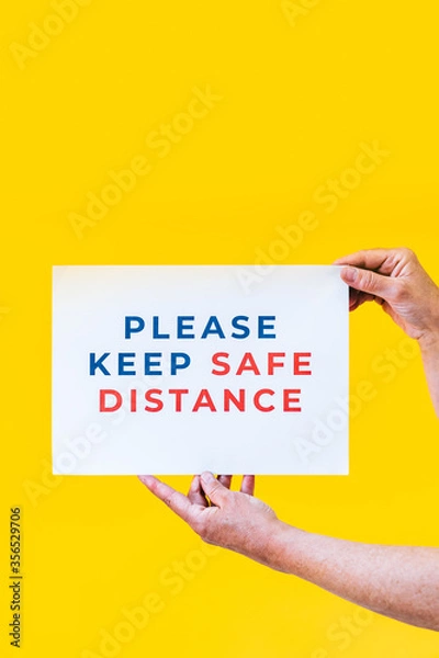 Fototapeta Please keep safe distance. Hands holding a sign for people to respect social distancing measures with an orange background.