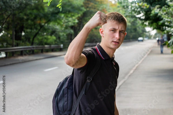 Fototapeta Portrait of a young man in a black T-shirt looking to camera with raising his hand with the clenched fist on the background of a road and green trees.