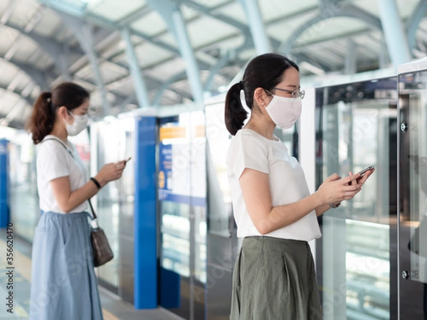 Obraz Two Asian women wearing medical face mask, using smartphone waiting for metro at train station platform, standing distance apart from other people.