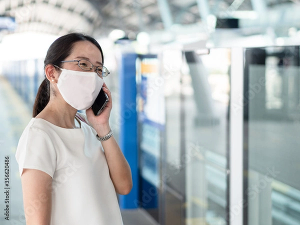 Obraz Beautiful Asian women wearing disposable medical face mask, using smartphone while waiting for metro at train station platform, as new normal trend and self-protection against Covid19 infection.