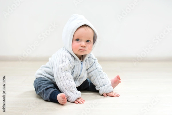 Fototapeta nine month baby sitting on floor and looking at camera, baby boy in hood at home sitting in front of white wall, place fot copy space