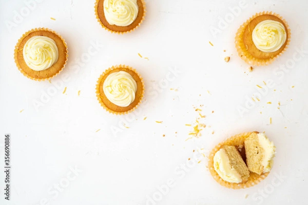 Fototapeta A top down view of five lemon cupcakes with vanilla frosting.  One cupcake has been cut in half to show centre and there are crumbs on the table.  Plain background flat lay style photo.