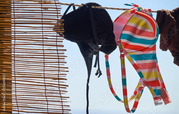 Fototapeta Colorful striped bikini hanging out to dry on a washing line in summer next to a reed curtain.