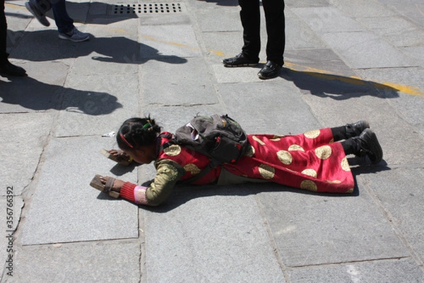 Fototapeta Little girl wearing traditional red cloathes with protection on hands praying in prostration in front of Jokhang temple in Lhasa on Barkhor square, Tibet