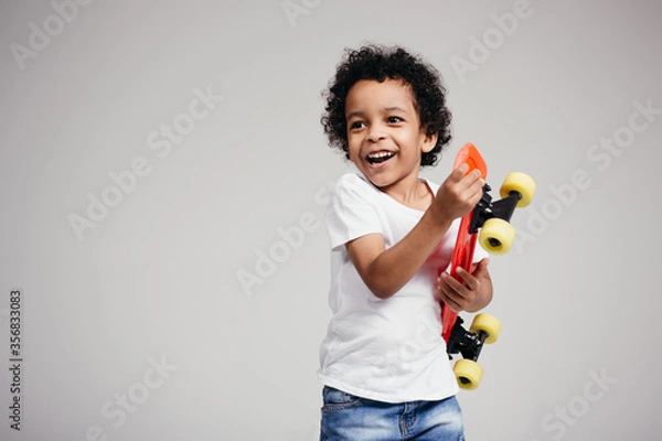 Fototapeta Smiling and laughing dark-skinned boy holds a red longboard in his hands and presses it to his chest on white background