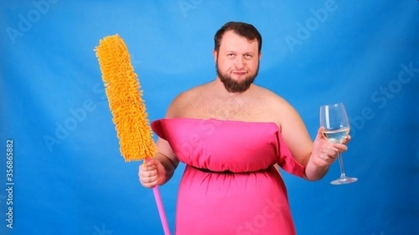 Fototapeta Funny bearded guy in a pink pillow dress with a mop and a glass of wine on a blue background. Crazy quarantine. Funny house cleaning. Fashion 2020. Put on a pillow. Challenge 2020 due to house