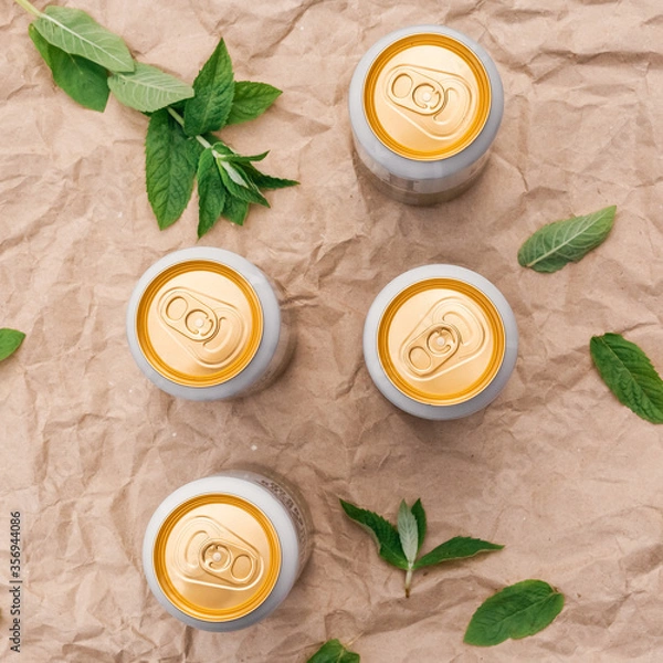 Fototapeta Four closed beer or soda gold aluminum cans, on a background of crumpled craft paper with mint leaves, top view, isolated, nobody. The concept of beer advertising.