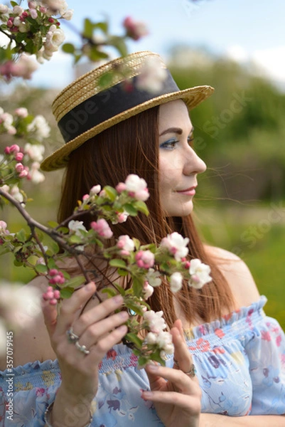 Fototapeta A beautiful girl in blue dress and straw hat holds flowering branch of Apple tree and looks into the distance in summer.