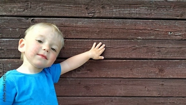 Fototapeta little smiling boy in a bright blue t-shirt shows something on the background of a brown wooden wall. Space for your advertising