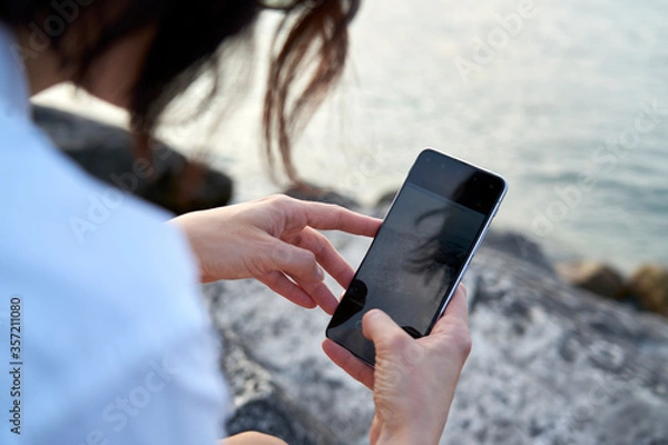 Fototapeta Close-up photograph of a blue screen smartphone held by a woman. In the background the sunset light and the sun reflecting on the blue water of Lake Garda.