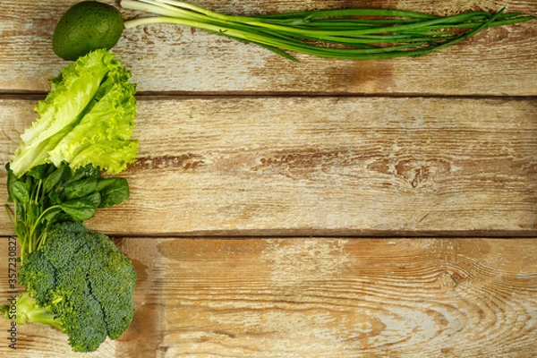 Fototapeta Green vegetables, broccoli, avocado, onions, spinach on the edge of a table of rough boards.