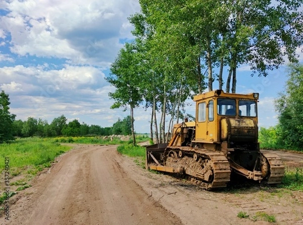 Fototapeta old yellow bulldozer stands on the side of a country road near trees against a blue sky with clouds on a sunny day