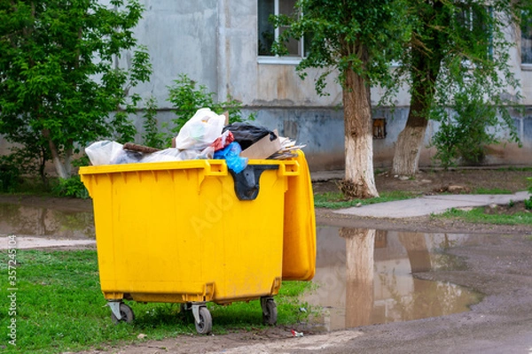Fototapeta Trash bin, garbage container full of waste. Yellow trash can near the apartment building. Environmental pollution concept, bad ecology.