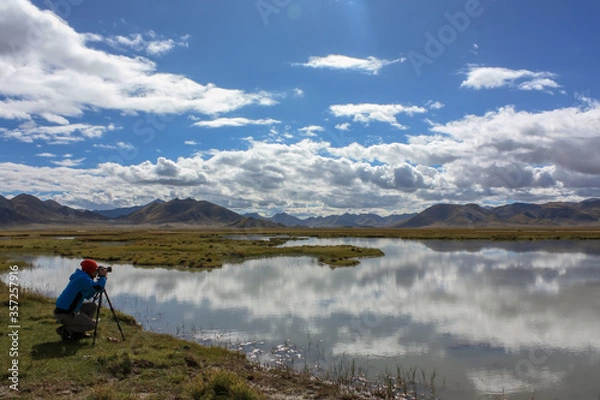 Fototapeta A tourist photographer setting up camera on tripod and taking a picture of beautiful reflection of hills and clouds in glass like waters of lake in Tibet, Asia