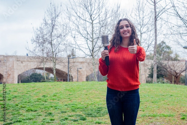 Fototapeta Portrait of pretty young woman model with rotating and styling brush  showing thumb up gesture, in the park, orange sweater and jeans, long curly hair. Place for your text in copy space.