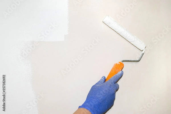 Obraz Repairs. A gloved hand holds a paint roller for painting the wall. Light streak. Real photo. Paint stained hand.