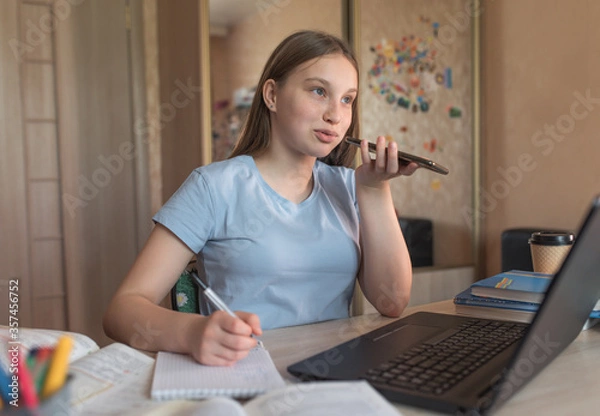 Fototapeta Teen girl doing homework, recording a voice message, making phone call, laptop monitor, e-education online lessons, distance learning. Social networks, watching a teacher’s lecture on Internet.