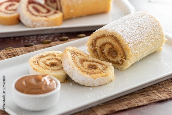 Fototapeta delicious roll cake with caramel dulce de leche and sugar sliced on white platter and blurred background