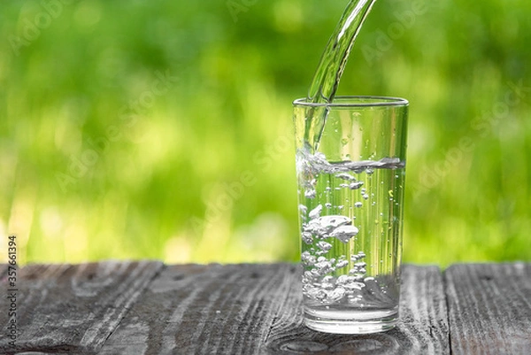 Fototapeta Water is poured into a glass against a background of greenery. A glass of water on a hot summer day with space to copy.