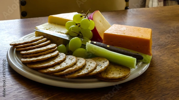 Fototapeta Rustic platter of crackers, knife and varied assortment of traditional cheeses, complemented with green grapes and celery. Set on an oak table. Interior image in natural light.