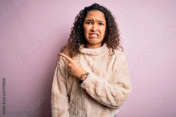 Fototapeta Young beautiful woman with curly hair wearing casual sweater standing over pink background Pointing aside worried and nervous with forefinger, concerned and surprised expression
