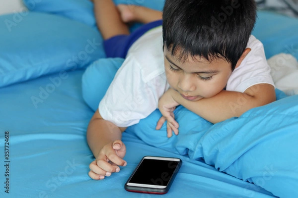 Obraz Cute asian boy playing games on smartphone. Child using a smartphone while lying on the bed. Social and technology concept.