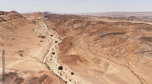 Fototapeta aerial view of wadi gevanim dotted with acacia trees looking west with the desert town of mitzpe ramon on the cliff in the right far background