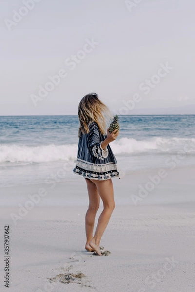Fototapeta Girl in ethnic dress on a white sand beach is dancing with fruit in her hands. Against the background of the ocean