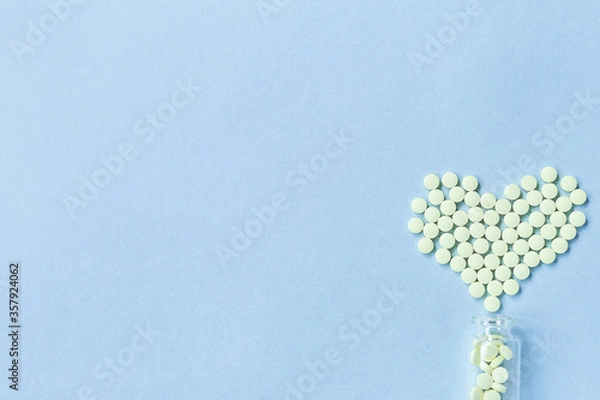 Fototapeta Group of yellow round tablets forms a heart shape on a blue background, spilled out of a glass jar. Selective focus. Concept of health care, treatment and prevention of diseases. Space for text.