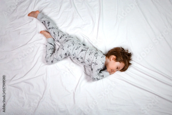 Fototapeta From above a boy is seen sleeping on a white bed in pajamas. Calm posture of the fetus. Concept of sleeping positions