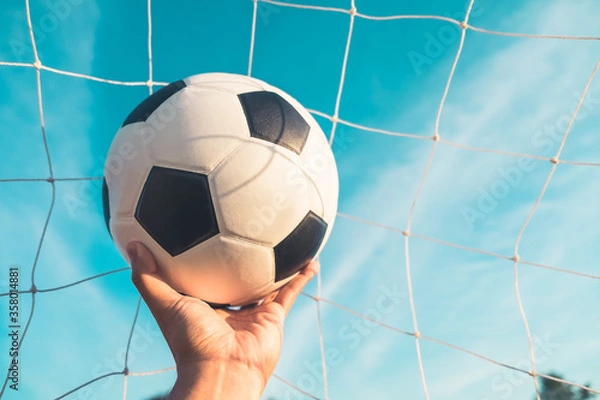Fototapeta Traditional soccer ball in goal net on blue sky nature background. A hand holding classic leather football equipment to play competitive game in stadium with copy space. Sport and exercise concept