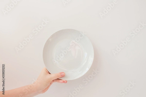 Fototapeta  Female hand holds a white round plate on a white background as a mockup with copy space. Ceramic flat saucer