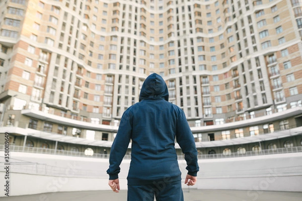Fototapeta Back of an athletic man in a hood and sportswear stands after training against the background of architecture and looks up. Strong muscular man stands with his back to the building.
