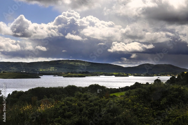 Fototapeta Vue panoramique sur un lac et ses îlots de terre, avec une ruine de tour médiévale en son centre. Ciel chargé et nuageux, collines, verdure et rochers.