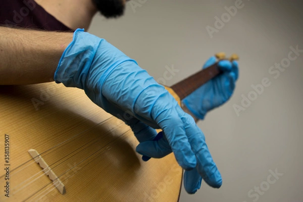 Fototapeta A man is playing a traditional string instrument called as 'baglama' in Turkish. He wears blue surgical gloves as a precation because of the covid-19 pandemic outbreak. New normal concept.