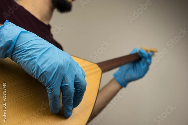 Fototapeta A man is playing a traditional string instrument called as 'baglama' in Turkish. He wears blue surgical gloves as a precation because of the covid-19 pandemic outbreak. New normal concept.