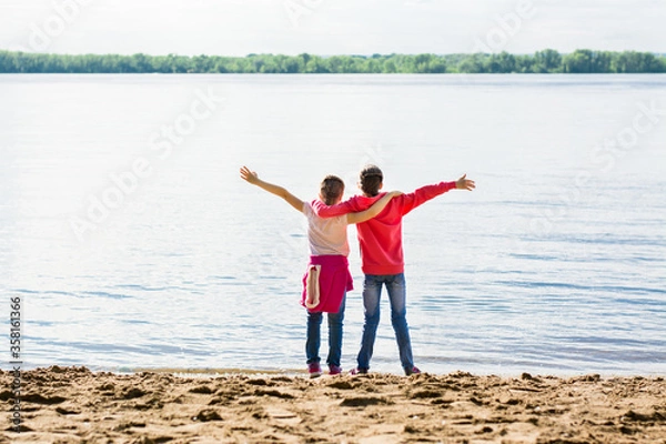 Fototapeta Local travel. Two girls hug on the river bank in the sand with arms outstretched. Summer tourism