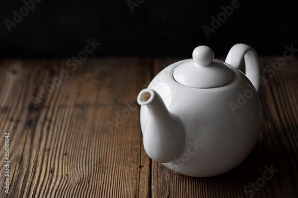 Fototapeta White ceramic mockup teapot for hot drinks on empty old wooden table near black wall in evening light with shadows. Horizontal with copy space for text and design. Rustic style.