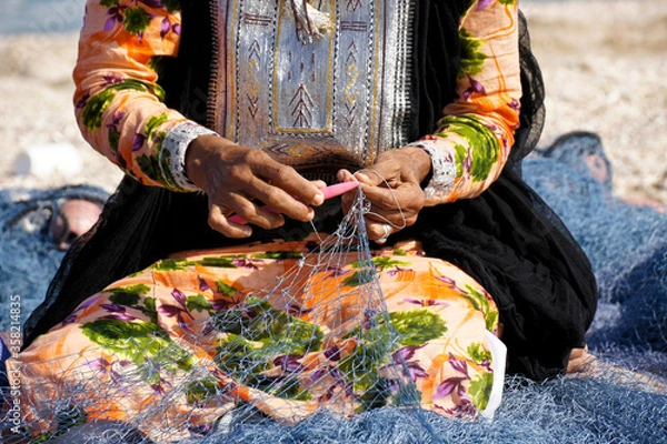 Fototapeta Omani woman in traditional dress mending fishing net on beach, Sur, Sultanate of Oman