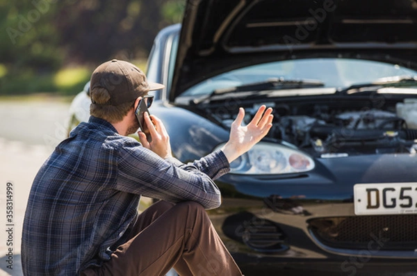 Obraz Young man next to his car, depressed and angry on the mobile phone telephone call after the car has broken down on the roadside waiting for assistance to arrive and recover or fix the vehicle